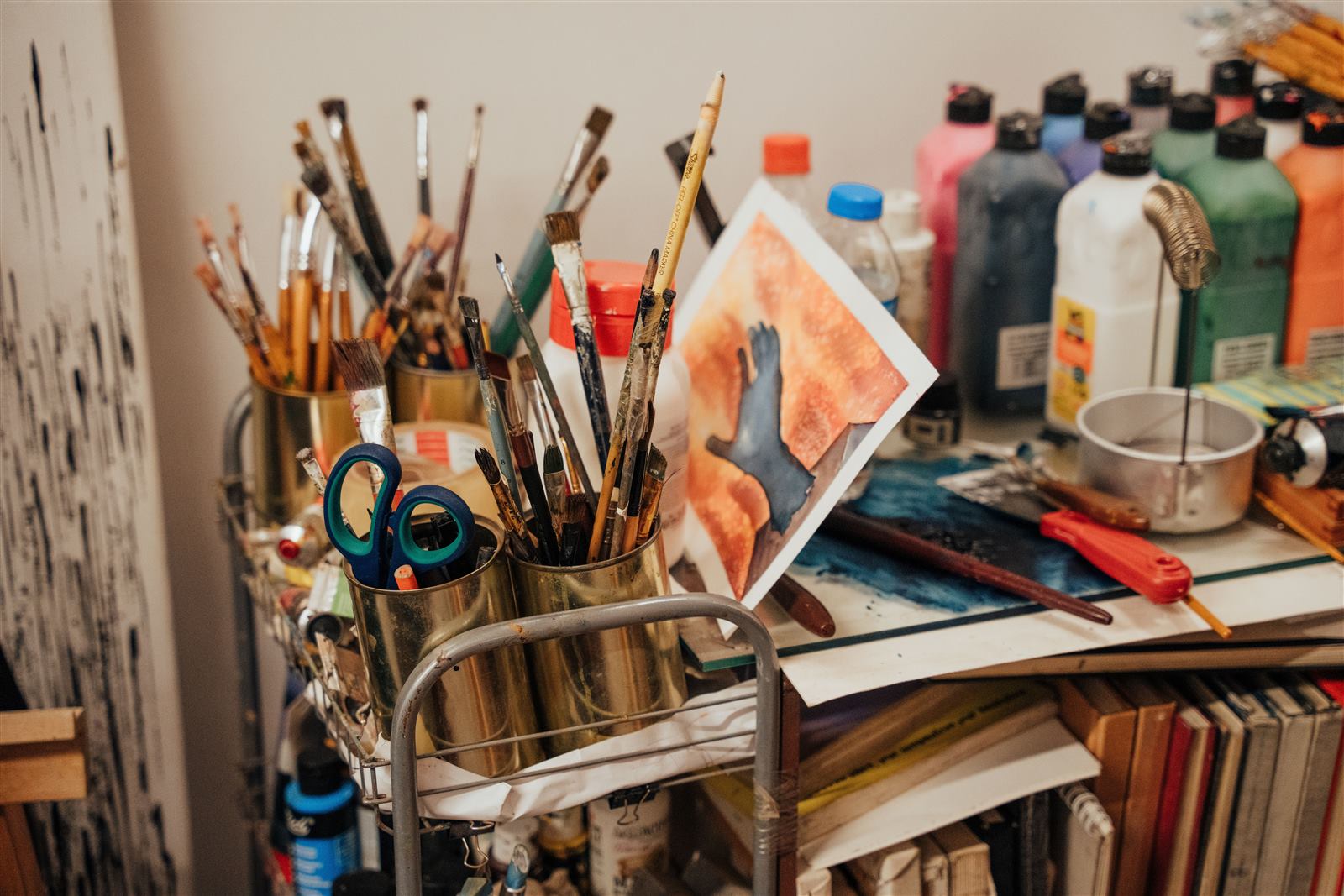 A craft workspace with paintbrushes in golden containers, colorful paint bottles, scissors and a watercolor painting